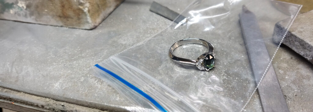 Shelby Engagment Ring Wide A silver ring with a dark green gemstone rests on a plastic ziplock bag on a workbench, surrounded by metal files and other tools—an Industrial Design NZ-inspired jewellery workshop with hints of CAD precision in every detail.