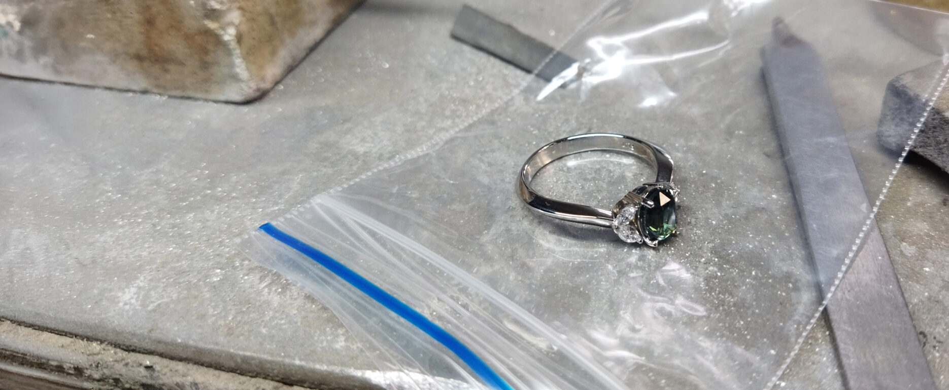 A silver ring with a dark green gemstone rests on a plastic ziplock bag on a workbench, surrounded by metal files and other tools—an Industrial Design NZ-inspired jewellery workshop with hints of CAD precision in every detail.