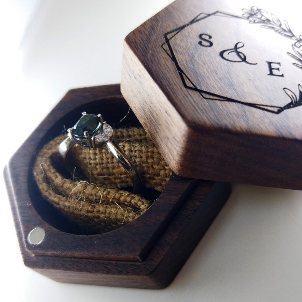 A silver ring with a dark green gemstone rests in a hexagonal wooden box, its lid engraved with floral designs and S & E initials—an elegant piece inspired by Industrial Design NZ, with the ring nestled on a hessian cushion inside.
