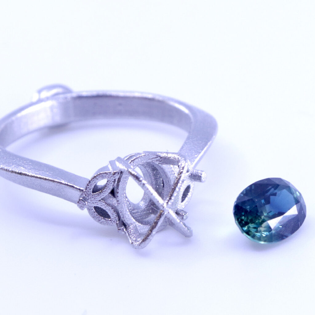 A silver ring setting sits beside a loose, oval-cut blue-green gemstone on a white surface, showcasing elegant Industrial Design NZ craftsmanship and waiting for the stone to be set into the ring.