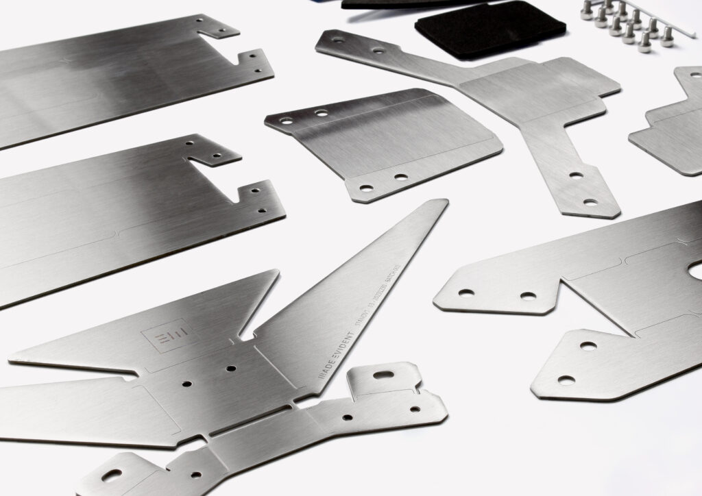 Several brushed metal plates and components with various cut-outs and shapes, likely designed using CAD, are laid out on a white surface alongside screws and small black rectangular pads.