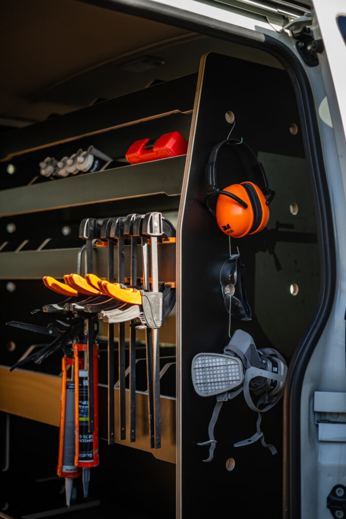 A neatly organised van interior, inspired by Industrial Design NZ principles, features shelves with tools, clamps, safety ear defenders, a respirator mask, caulking guns, and tubes of sealant. The setup is efficient and ready for work.