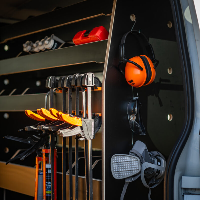 A neatly organised van interior, inspired by Industrial Design NZ principles, features shelves with tools, clamps, safety ear defenders, a respirator mask, caulking guns, and tubes of sealant. The setup is efficient and ready for work.