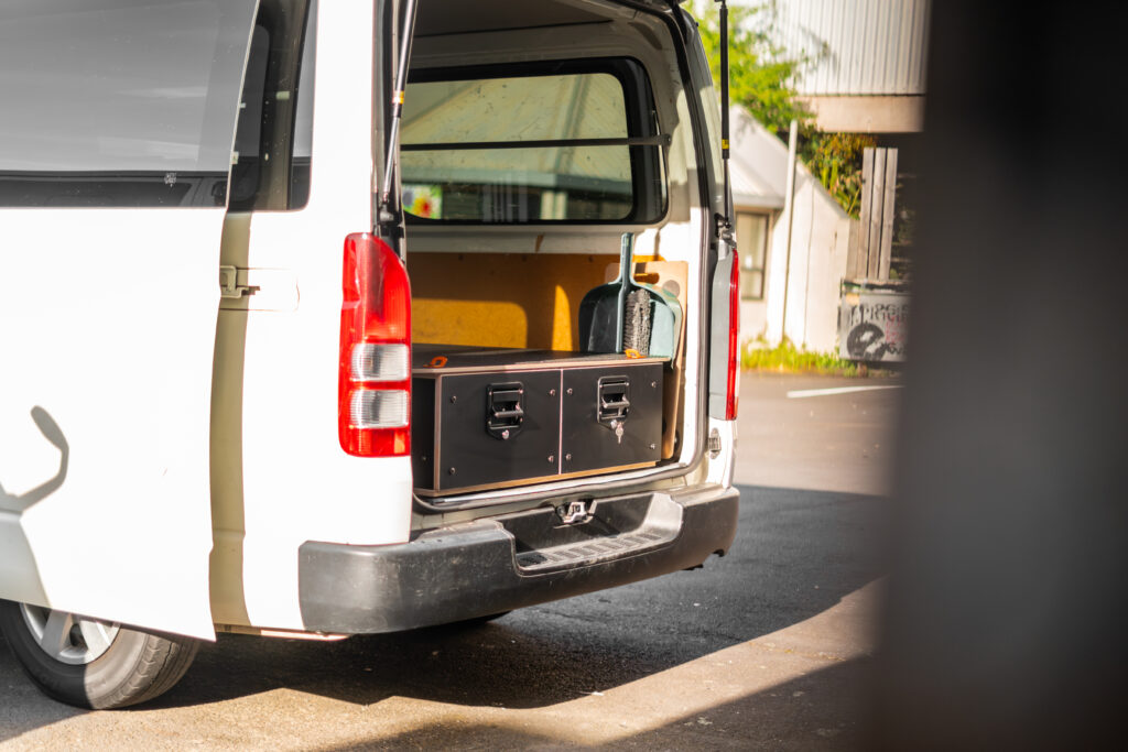 A white van with its rear door open reveals a large black storage box inside—an example of Industrial Design NZ. The van is parked outdoors on a sunny day, and part of a building is visible in the background.