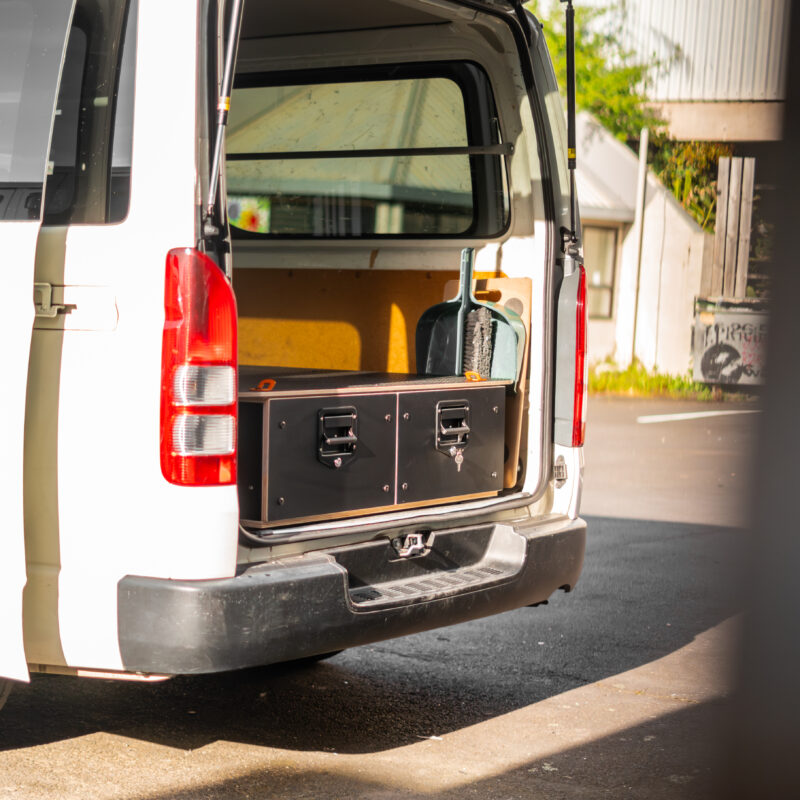 A white van with its rear door open reveals a large black storage box inside—an example of Industrial Design NZ. The van is parked outdoors on a sunny day, and part of a building is visible in the background.