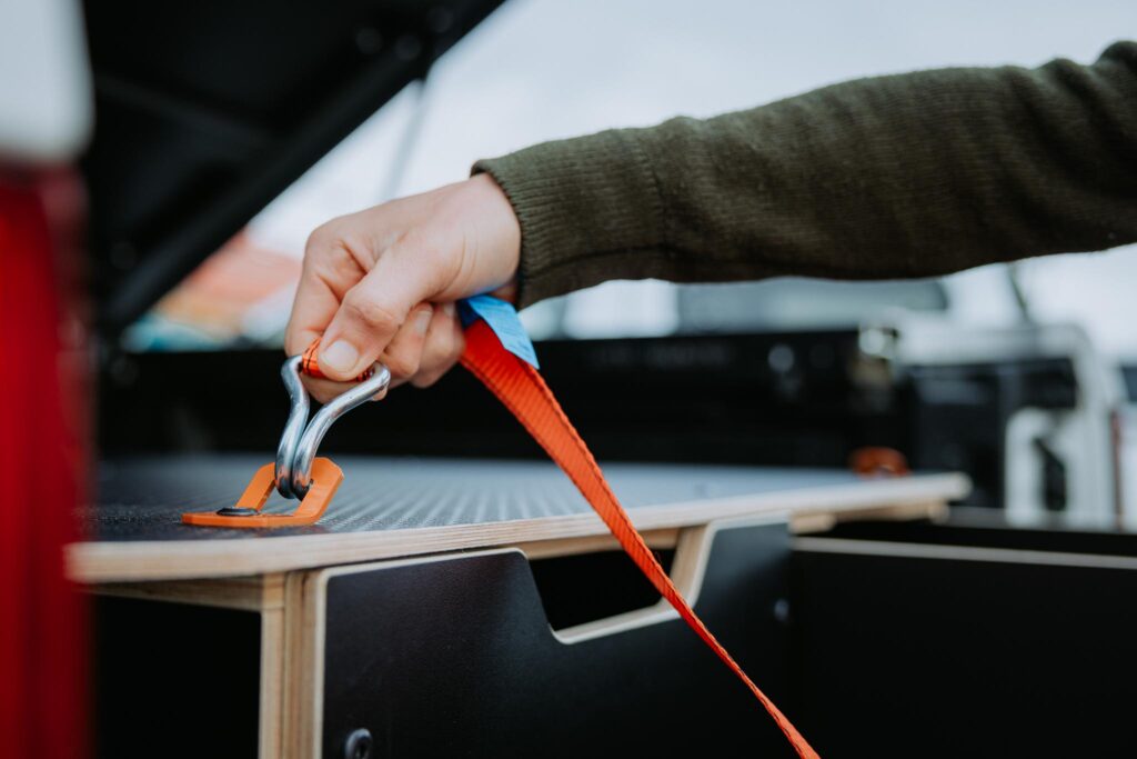 A person’s hand secures an orange tie-down strap with a metal hook onto a catch inside the back of a vehicle with a storage compartment, demonstrating attention to detail in Industrial Design NZ.