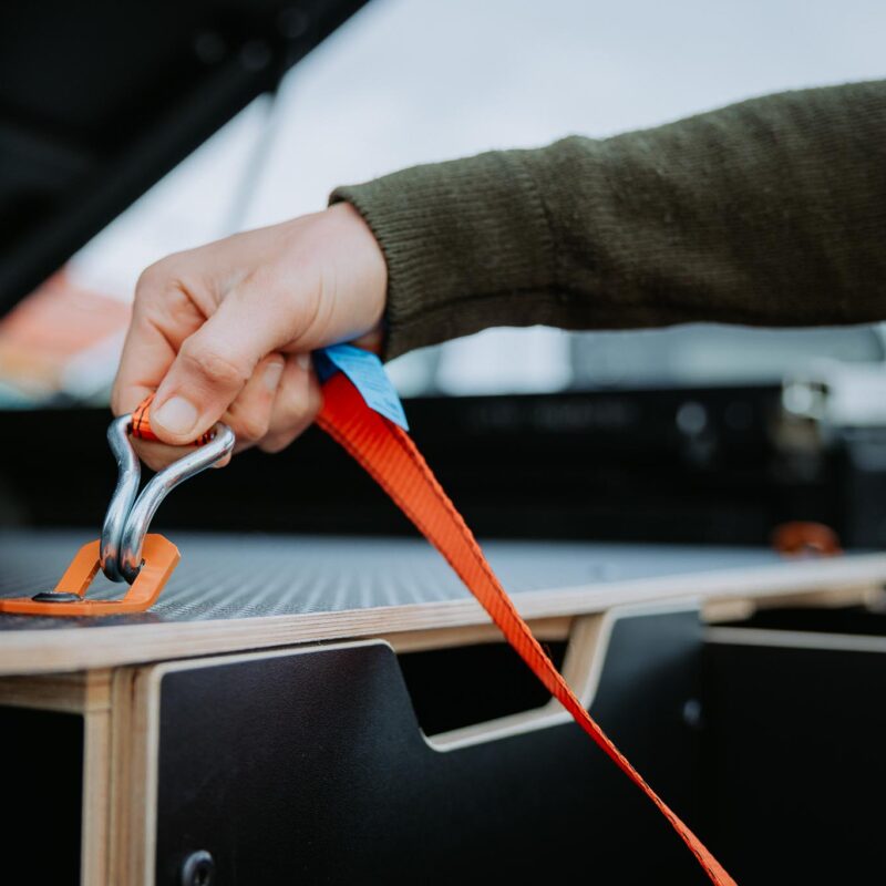 A person’s hand secures an orange tie-down strap with a metal hook onto a catch inside the back of a vehicle with a storage compartment, demonstrating attention to detail in Industrial Design NZ.