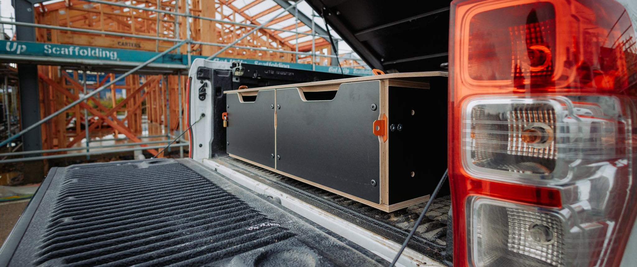 A pickup truck with its tailgate down shows a black and wood storage drawer system, reflecting expert Product Design NZ. In the background, a building under construction with scaffolding is visible.