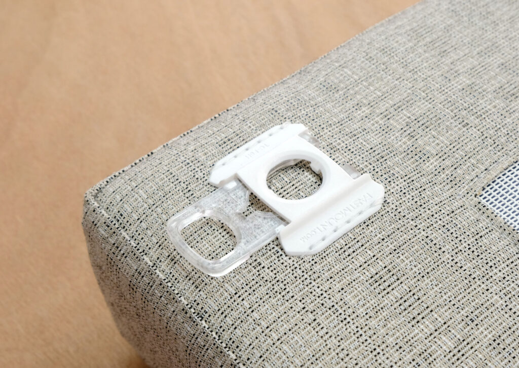 A white and clear plastic bread bag clip, designed by a freelance industrial designer, rests on a light grey, textured fabric surface atop a wooden table.