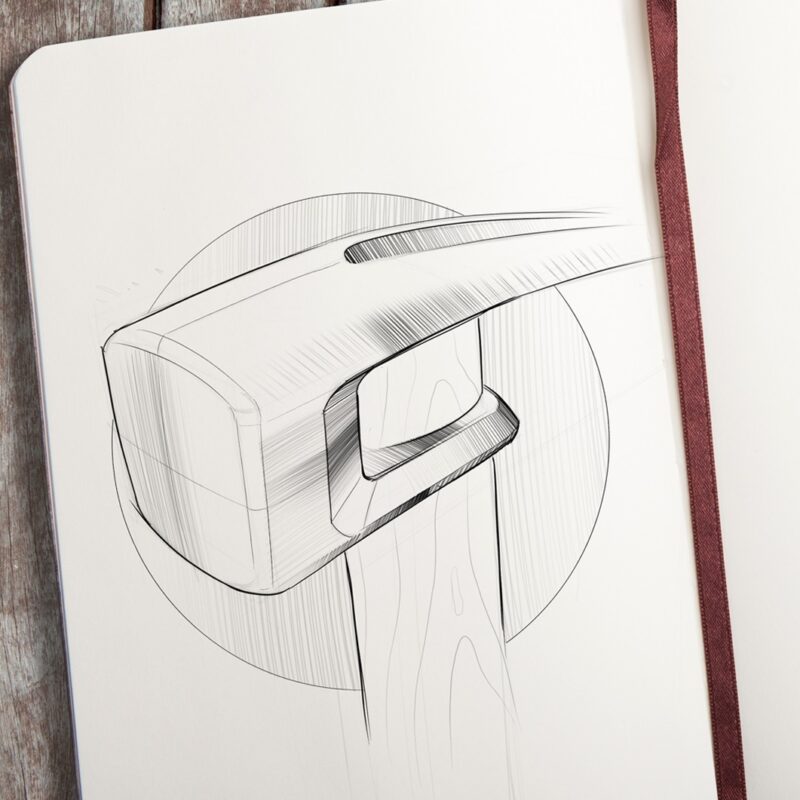 A sketch by Evan Thomas of a sledgehammer head with a wooden handle, drawn in black ink on white paper in a notebook, rests on a wooden surface with a brown ribbon bookmark visible—showcasing Industrial Design Auckland flair.