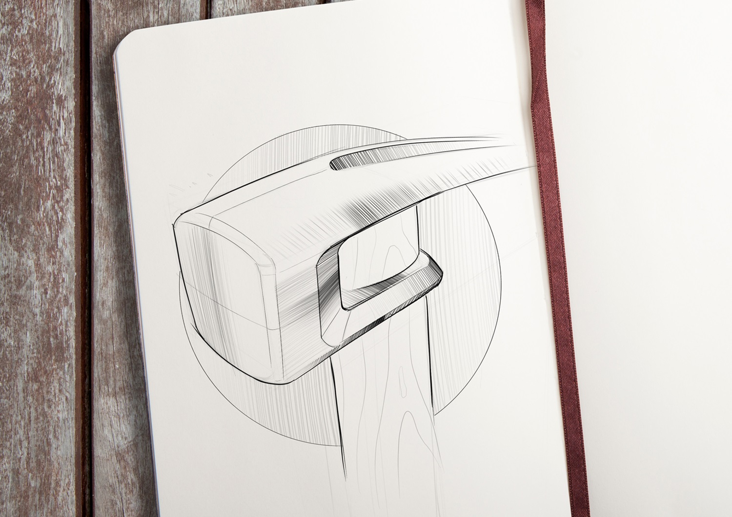A sketch by Evan Thomas of a sledgehammer head with a wooden handle, drawn in black ink on white paper in a notebook, rests on a wooden surface with a brown ribbon bookmark visible—showcasing Industrial Design Auckland flair.