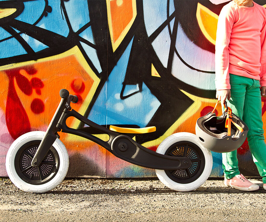 A child in a pink jumper and green trousers stands next to a black balance bike with white tyres, holding a grey helmet, in front of a colourful graffiti wall—an inspiring scene for any Product Design NZ enthusiast.