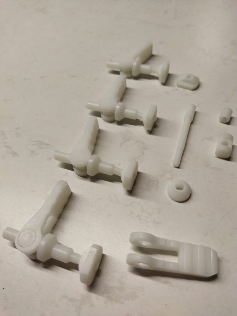 Several small, white plastic mechanical parts are arranged on a light-coloured surface. These components, reflecting principles of Design for Manufacture, include joints, rods, and circular pieces—possibly for assembly into a larger object.
