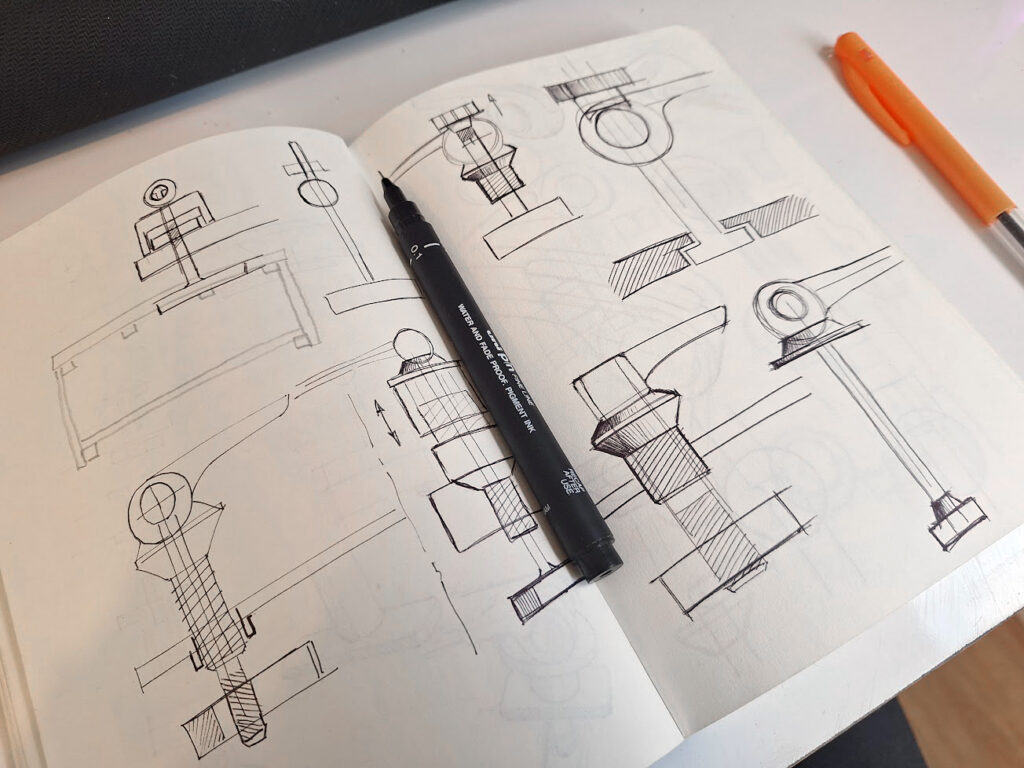 An open sketchbook displays technical pen sketches of mechanical parts and cross-sections, reflecting the creative process of a Freelance Industrial designer. A black pen lies across the pages, and an orange pen rests nearby on a white desk.