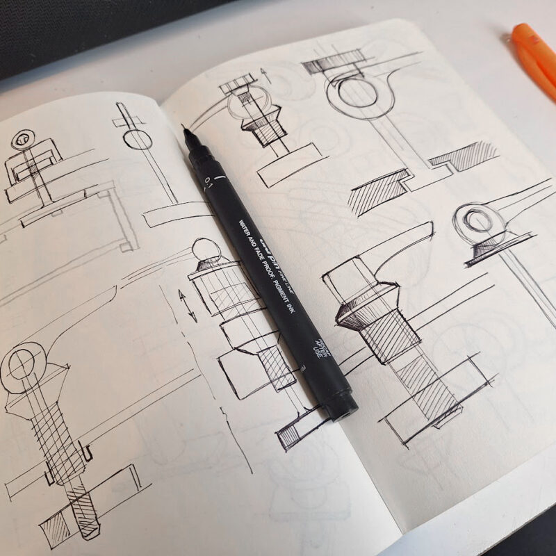 An open sketchbook displays technical pen sketches of mechanical parts and cross-sections, reflecting the creative process of a Freelance Industrial designer. A black pen lies across the pages, and an orange pen rests nearby on a white desk.