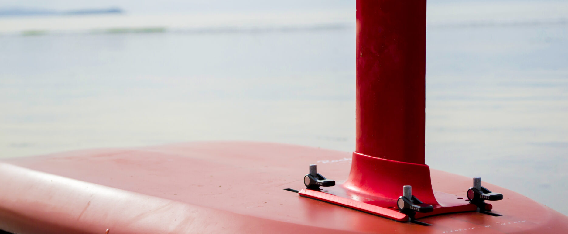 Close-up of a red hydrofoil surfboard resting on calm water, showing the base of the mast and smooth board surface with F-one written in white lettering. Product Design NZ expertise influences its sleek, CAD-enhanced lines. Out-of-focus shoreline and sky behind.