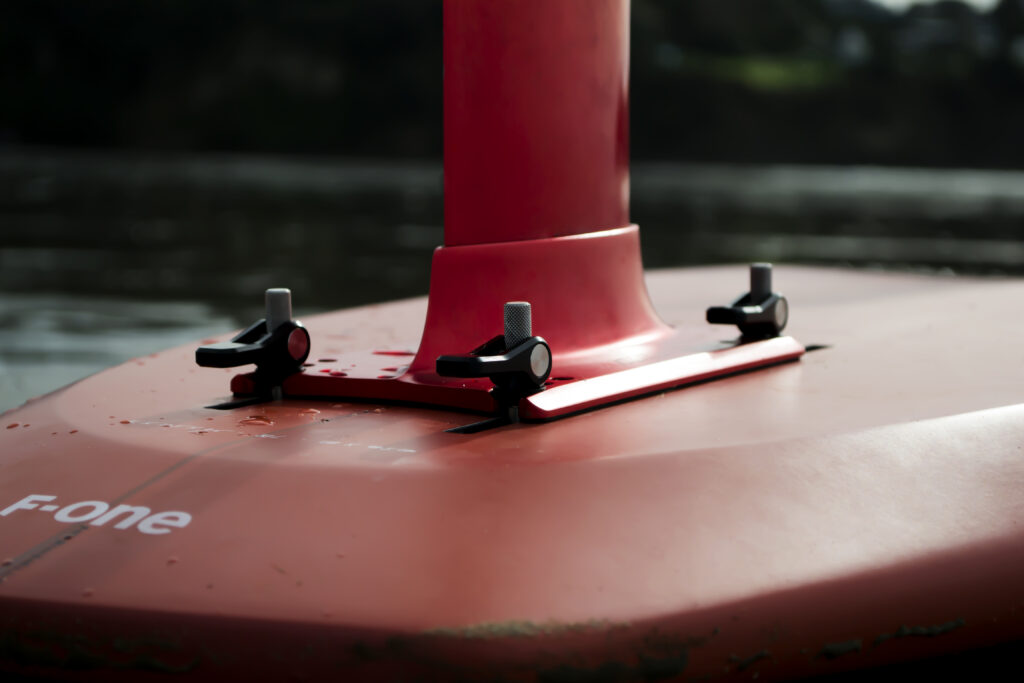 Close-up of a red hydrofoil mast base, crafted with precision Product Design NZ expertise, attached to a board with four black screws and wing nuts. Water droplets highlight the F-one branding on the smooth surface.