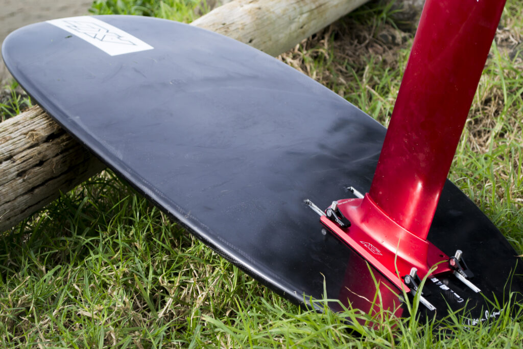 A black hydrofoil surfboard with a red foil mast, designed for manufacture by a freelance industrial designer, lies on green grass next to a wooden log, ready for use.