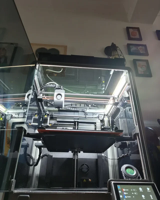 A 3D printer is in operation inside a glass enclosure, illuminated by bright LED strips. Framed pictures and other items are visible on the wall, reflecting the workspace of a freelance industrial designer skilled in CAD and Design for Manufacture. A display screen is visible below.