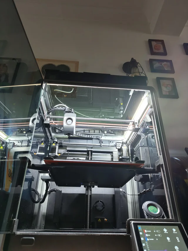 A 3D printer is in operation inside a glass enclosure, illuminated by bright LED strips. Framed pictures and other items are visible on the wall, reflecting the workspace of a freelance industrial designer skilled in CAD and Design for Manufacture. A display screen is visible below.